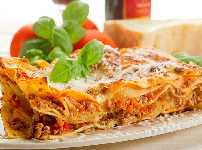 Close-up of a layered lasagna dish garnished with basil leaves and grated cheese, with tomatoes and bread in the background