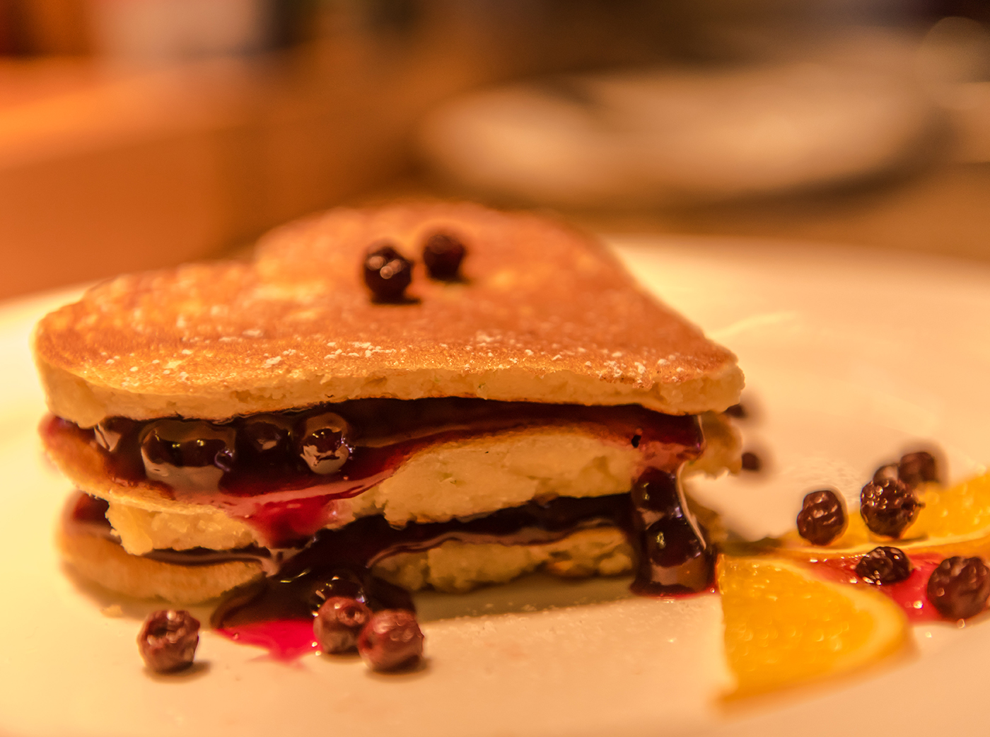 Close-up of a stacked pancake dessert with blueberries, berry sauce, and orange slice garnish on a white plate