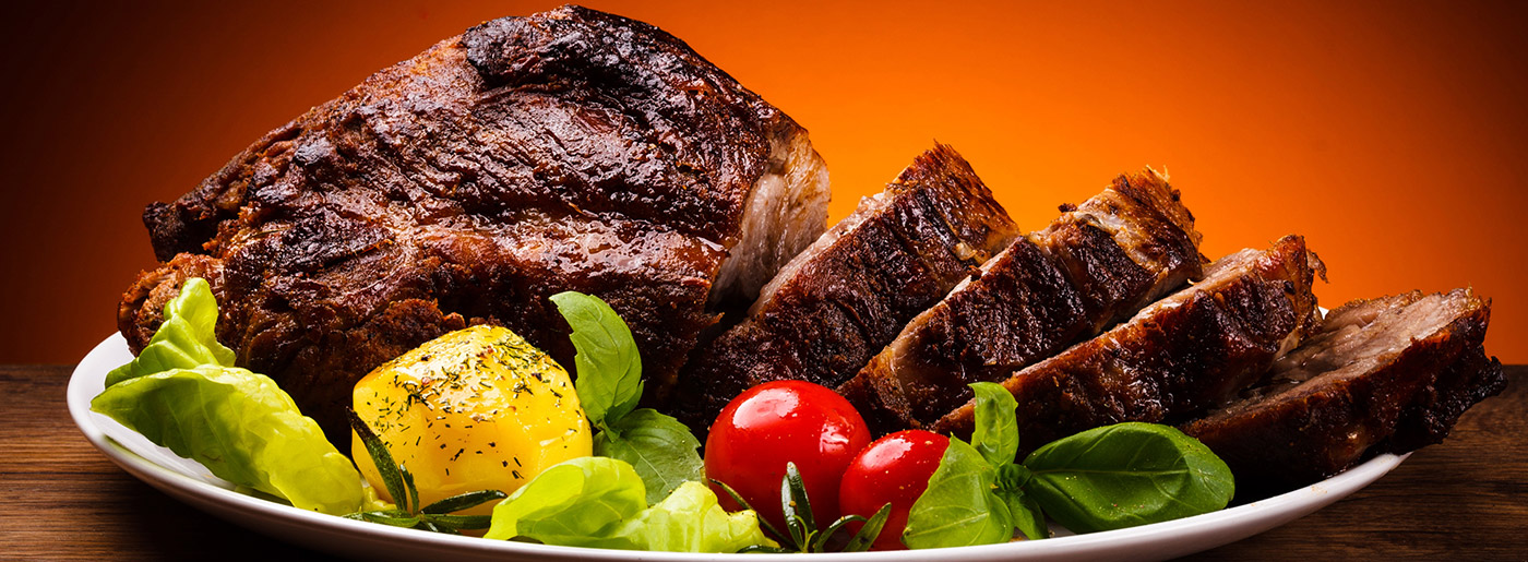 Plated roasted meat with sliced ribs, cherry tomatoes, lettuce, and seasoned corn on a white plate against an orange background
