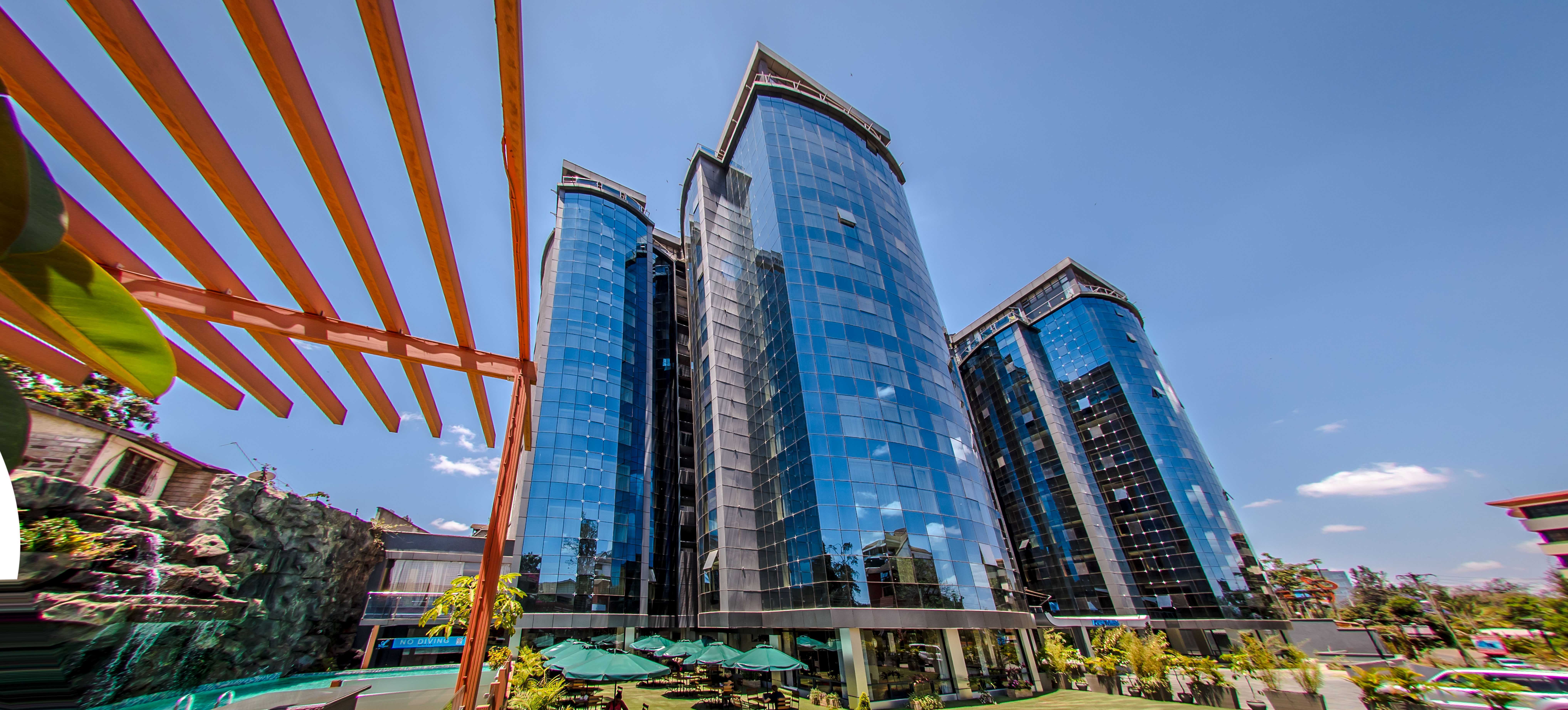 Modern glass-clad high-rise towers under a blue sky, seen from below with an orange pergola structure in the foreground