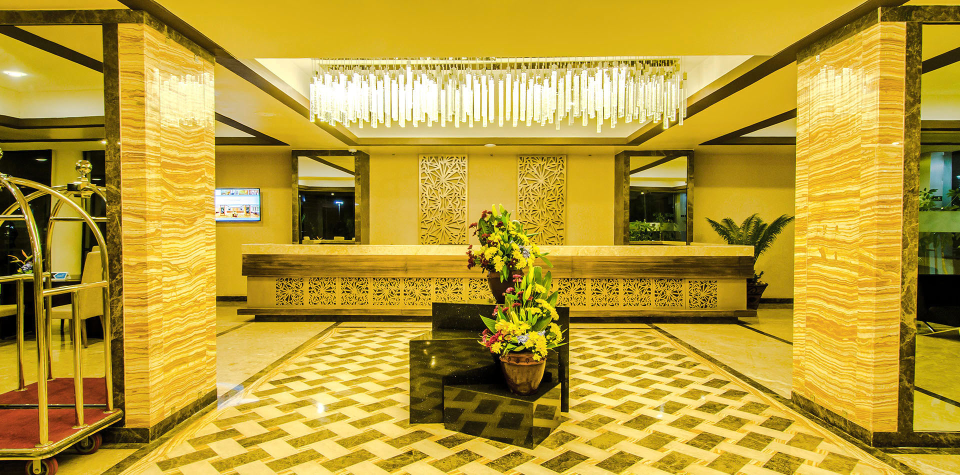Hotel lobby reception area with patterned marble floor, gold-toned lighting, decorative wall panels, and a chandelier