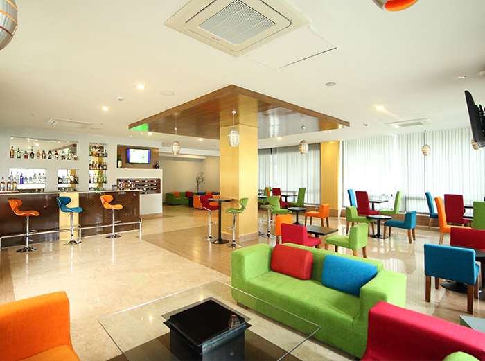Colorful lounge and bar area with mismatched red, green, orange, and blue seating, a stocked bar counter, and modern ceiling lighting